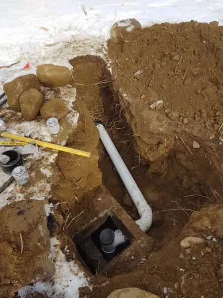 Excavated trench in snowy ground showing a white PVC pipe connected to an underground black pipe fitting.
