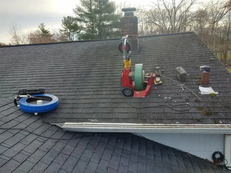 Roof with a chimney, a red machine with a green wheel labeled 'Spartan 301', a blue reel, and a black toolbox, with leafless trees in the background.
