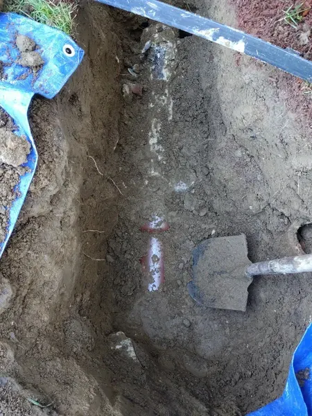 Shovel resting in a rectangular excavation revealing a partially exposed underground pipe wrapped with patches of red tape.