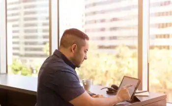 Man sitting at a table using a digital tablet with a cityscape visible through large windows behind him.