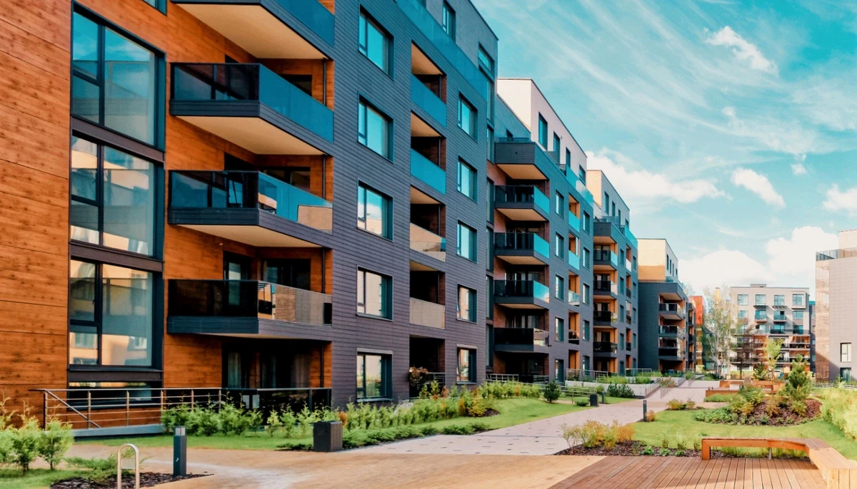 Modern apartment complex with multiple balconies overlooking landscaped garden and walking paths under a partly cloudy sky.