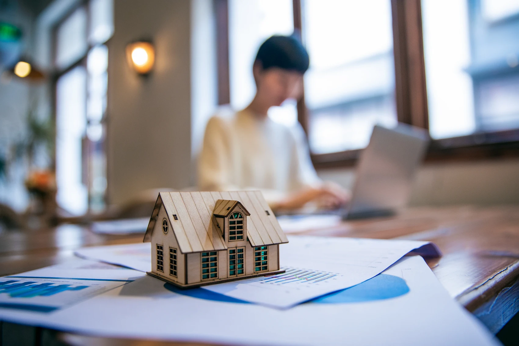 Wooden model house on financial documents with a person working on a laptop in the background.