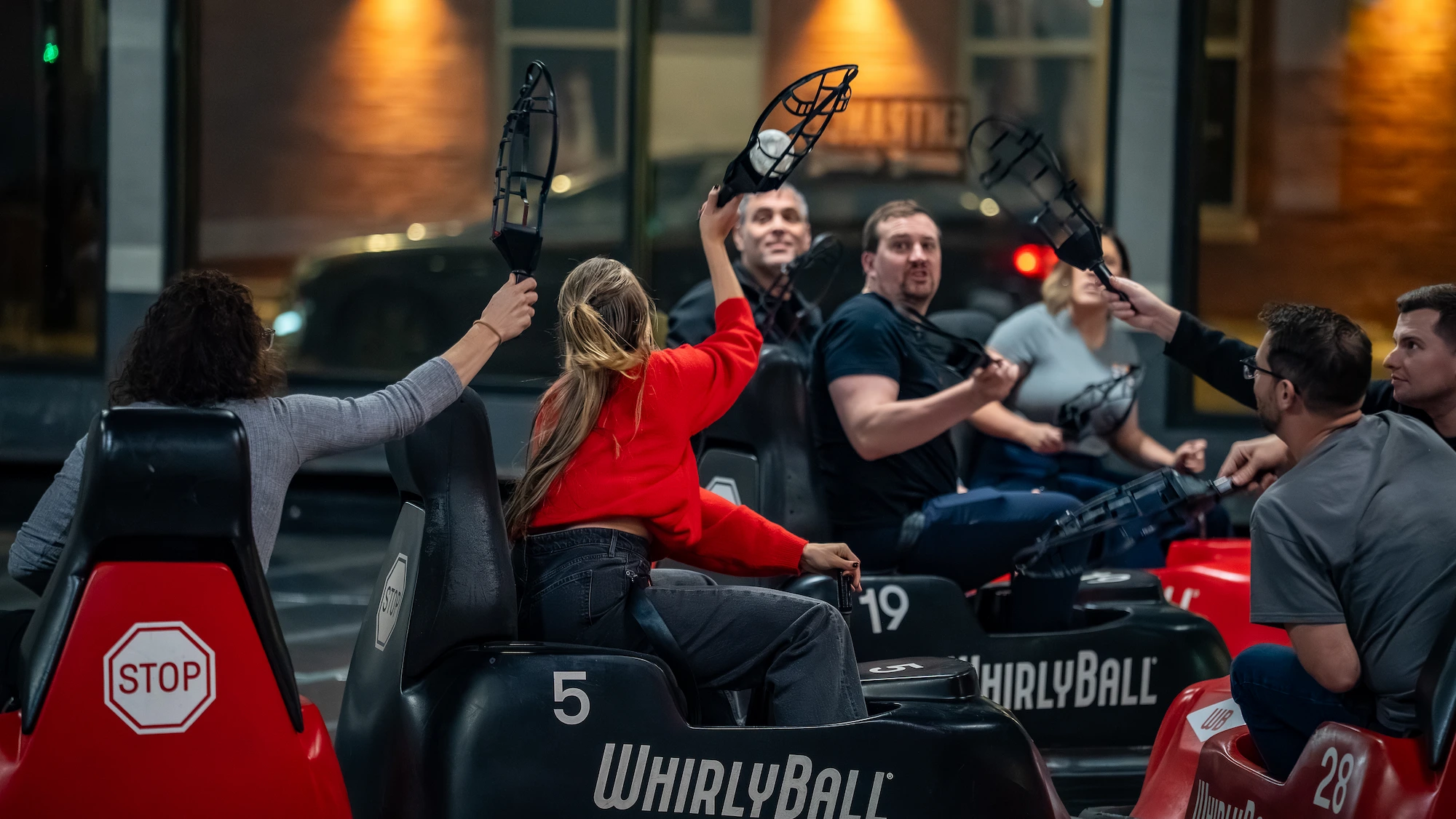 Group of adults playing WhirlyBall indoors, sitting in numbered cars and raising paddles.