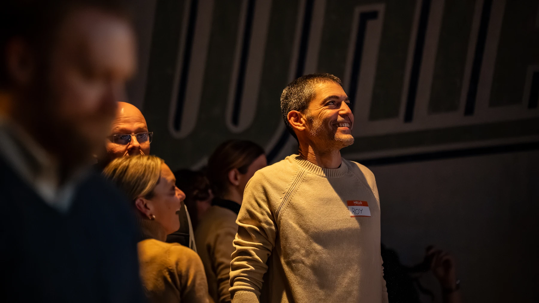 Man smiling and standing in a dimly lit room with several other people.