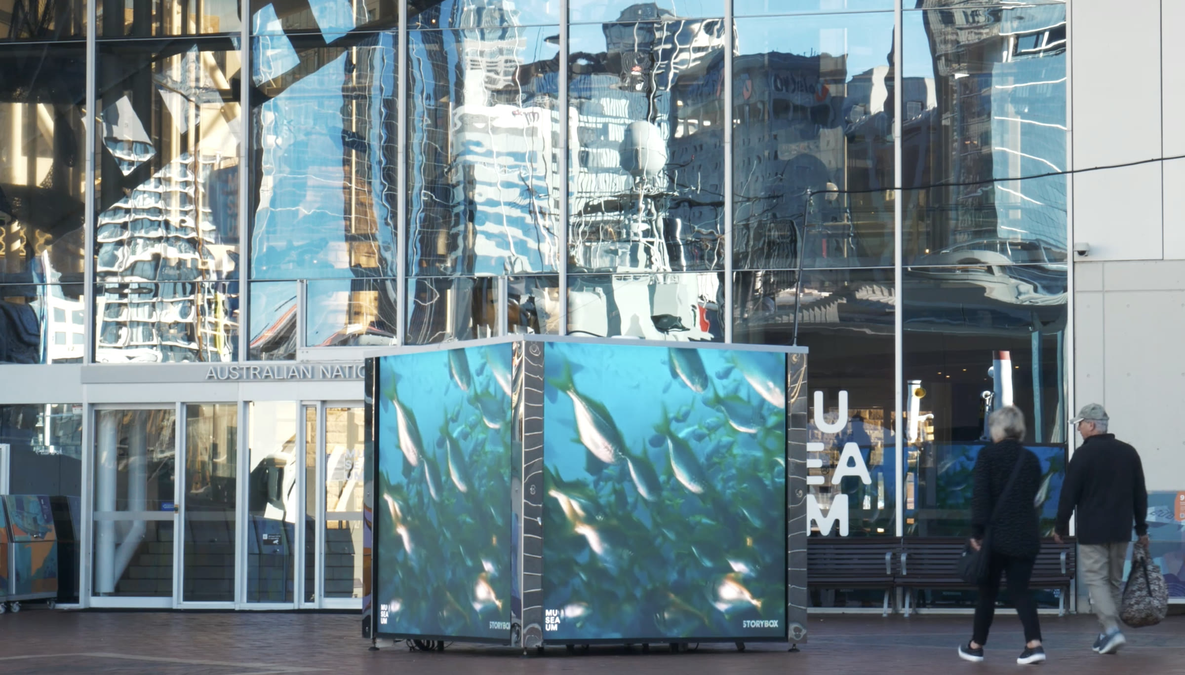 Visitors passing the STORYBOX cube at the entrance to the Australian National Maritime Museum, the screens showing a school of fish.