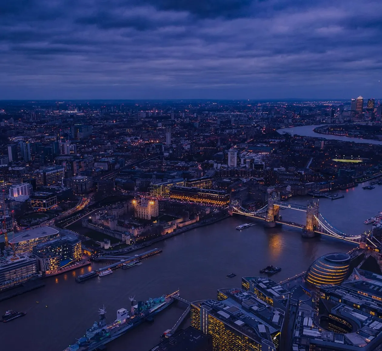 Aerial view of London at dusk showing Tower Bridge, the River Thames, and city buildings illuminated under a cloudy sky.