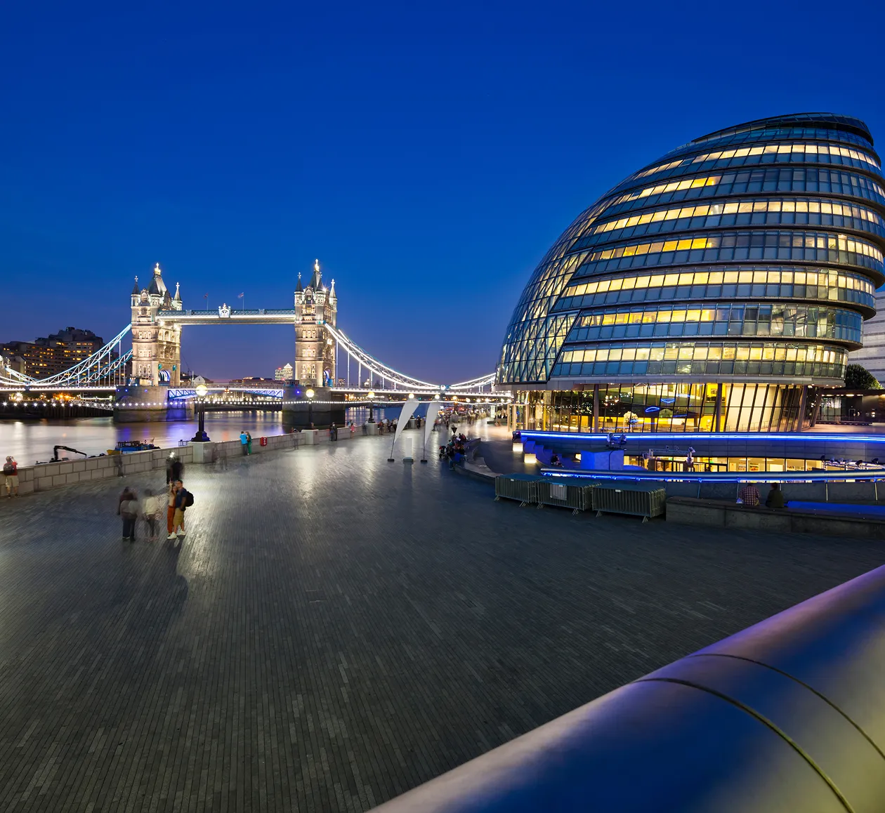 Illuminated Tower Bridge and modern glass City Hall building along the River Thames at dusk in London.