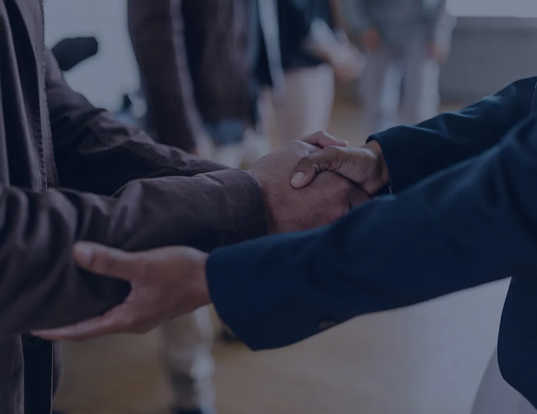Two people shaking hands with one person’s other hand supporting the handshake in a blurred indoor setting.