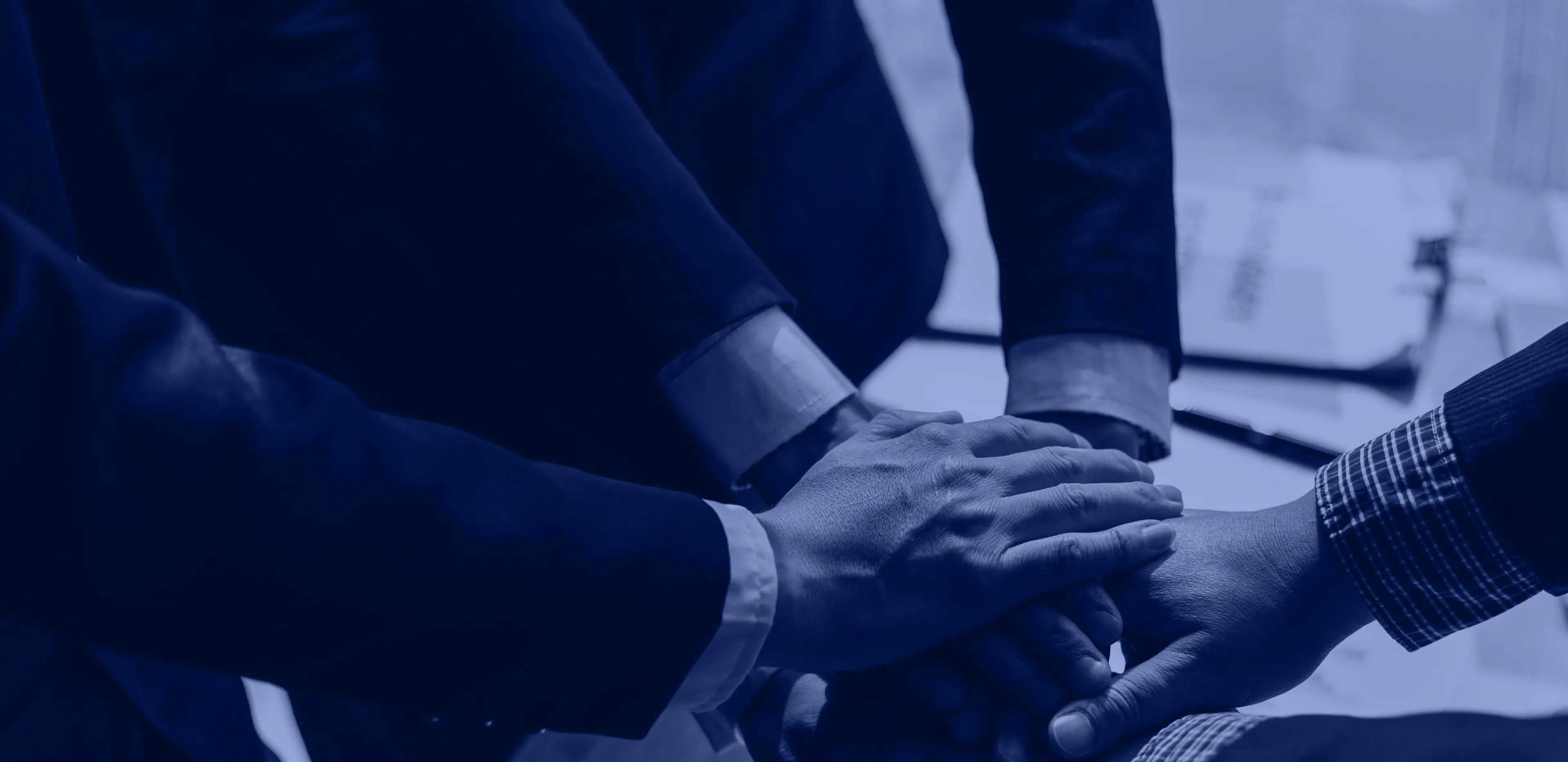 Four people in business attire stacking their hands together in a teamwork gesture over a desk with documents.