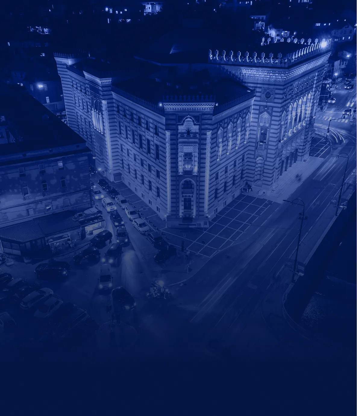 Illuminated large historic building at night with surrounding streets and parked cars.