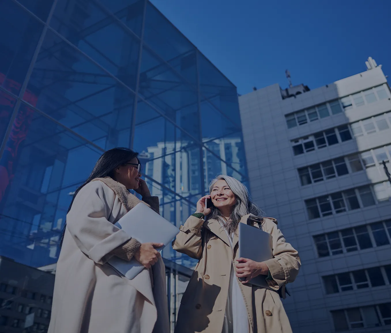 Two professional women in trench coats holding laptops and talking on phones outside modern office buildings.