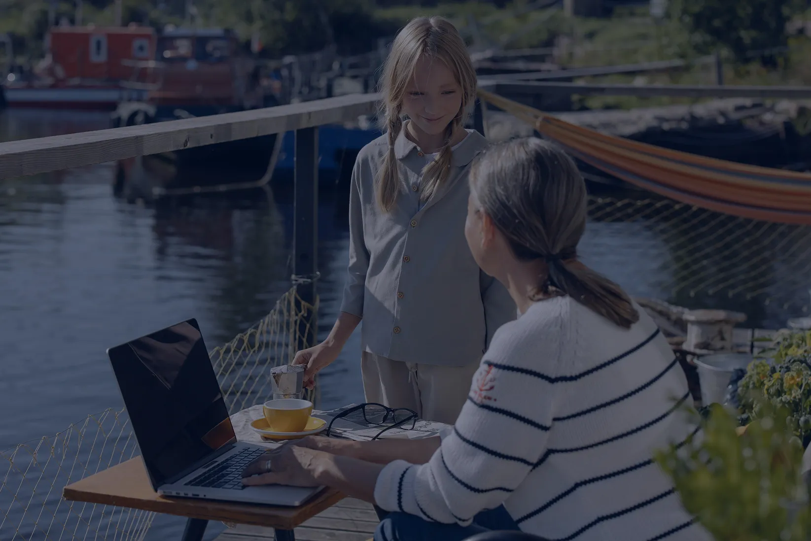Young girl standing by a woman sitting with a laptop on a dock by the water, with a yellow cup and glasses on the table.