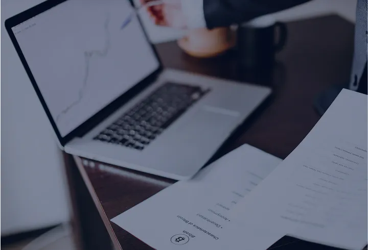 Person holding financial documents next to a laptop displaying a line graph on a wooden desk.