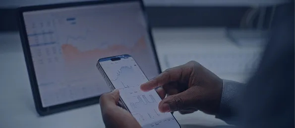 Person holding a smartphone displaying financial charts, with a tablet showing graphs in the background.