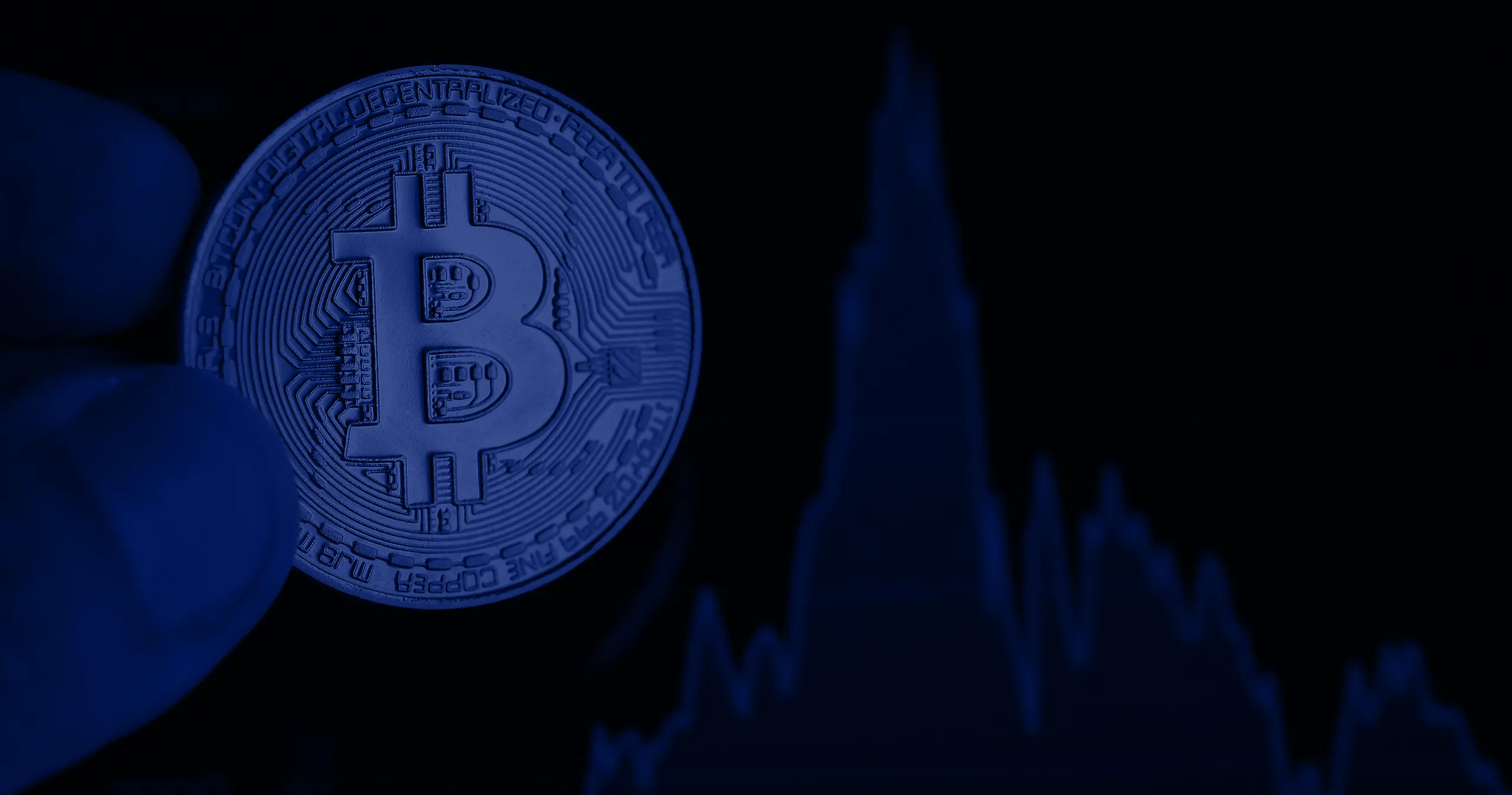 Close-up of a hand holding a physical Bitcoin coin with a blurred cryptocurrency price chart in the background.