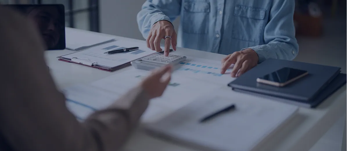 Person using a calculator at a desk with documents, pens, and a smartphone nearby, during a discussion with another individual.