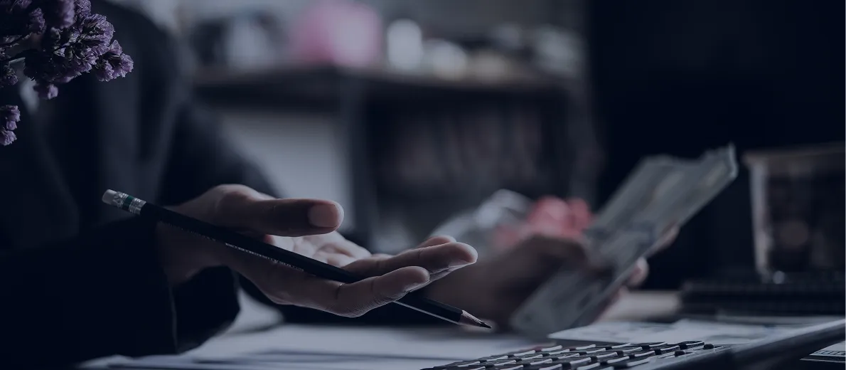 Person holding a pencil and counting money with a calculator on the desk.