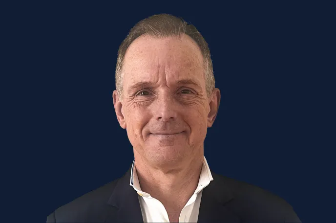 Middle-aged man with short hair wearing a dark blazer and white shirt smiling against a dark blue background.