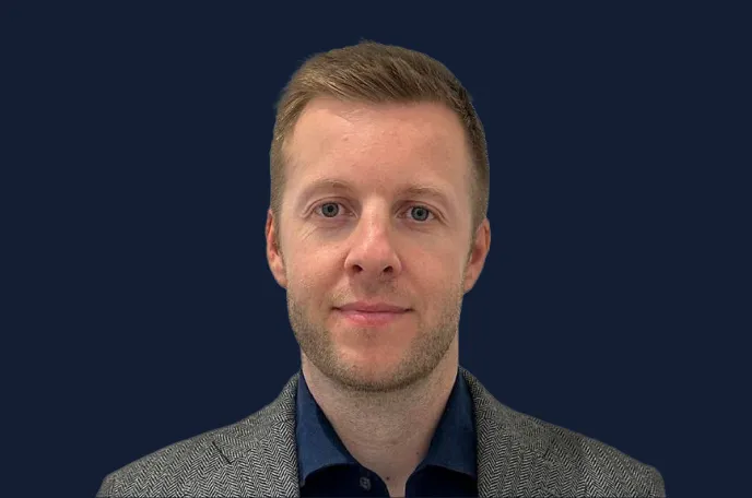 Portrait of a man with short light brown hair wearing a gray herringbone jacket and dark blue shirt against a dark blue background.