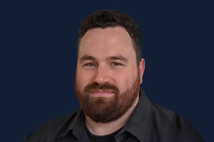 Portrait of a man with dark curly hair, beard, and mustache wearing a dark shirt against a dark blue background.