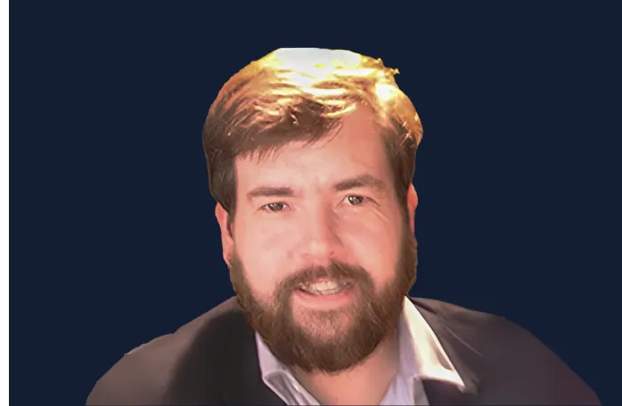 Smiling man with brown hair and beard wearing a suit jacket and light-colored shirt against a dark blue background.