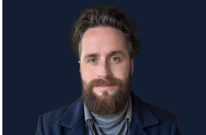 Bearded man with dark hair wearing a dark jacket and gray turtleneck, looking at the camera against a dark blue background.