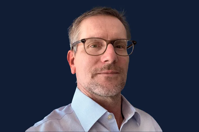 Middle-aged man with short hair, glasses, and light beard wearing a light blue button-up shirt against a dark blue background.