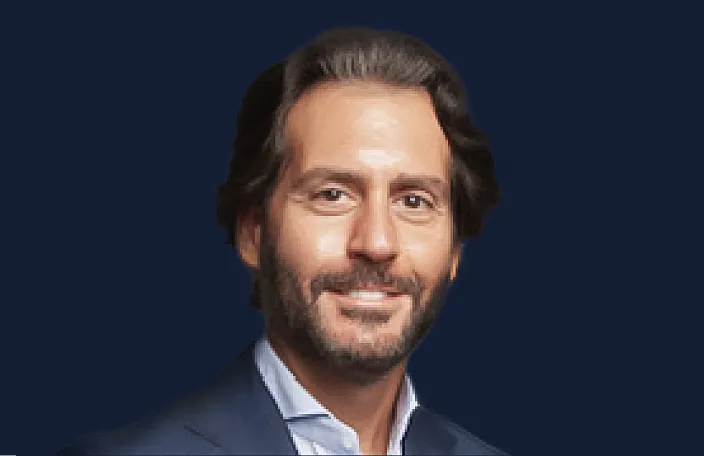 Headshot of a man with dark hair and beard, wearing a navy blazer and light blue shirt against a dark blue background.