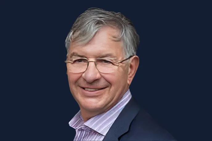 Smiling older man with gray hair wearing glasses, a striped shirt, and a dark blazer against a navy blue background.