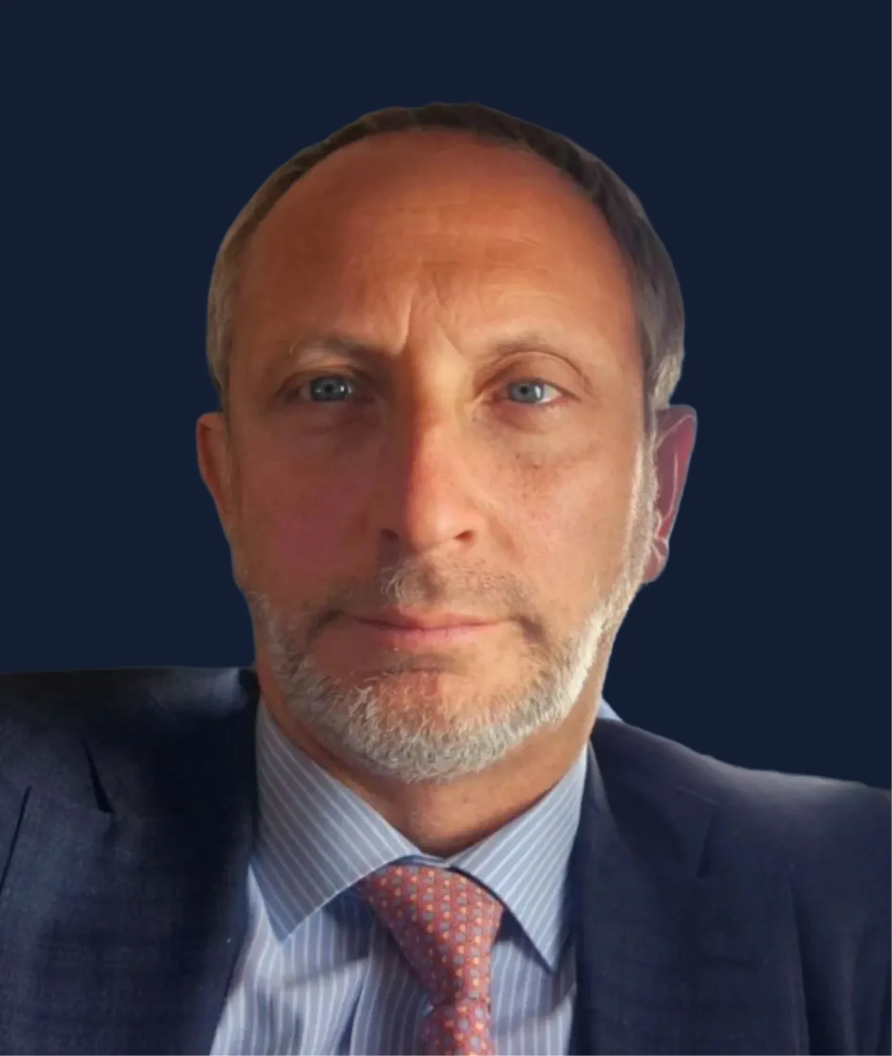 Close-up portrait of a middle-aged man with light beard, blue eyes, wearing a suit, striped shirt, and patterned tie against a dark blue background.