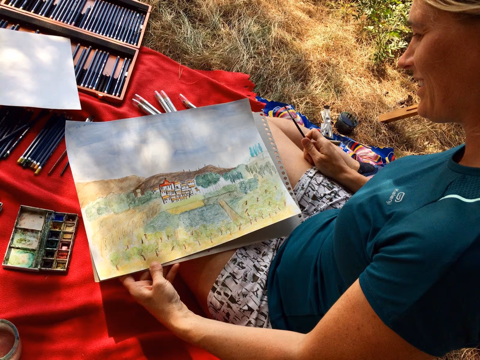 A woman is sitting on a blanket and drawing a picture of a house.