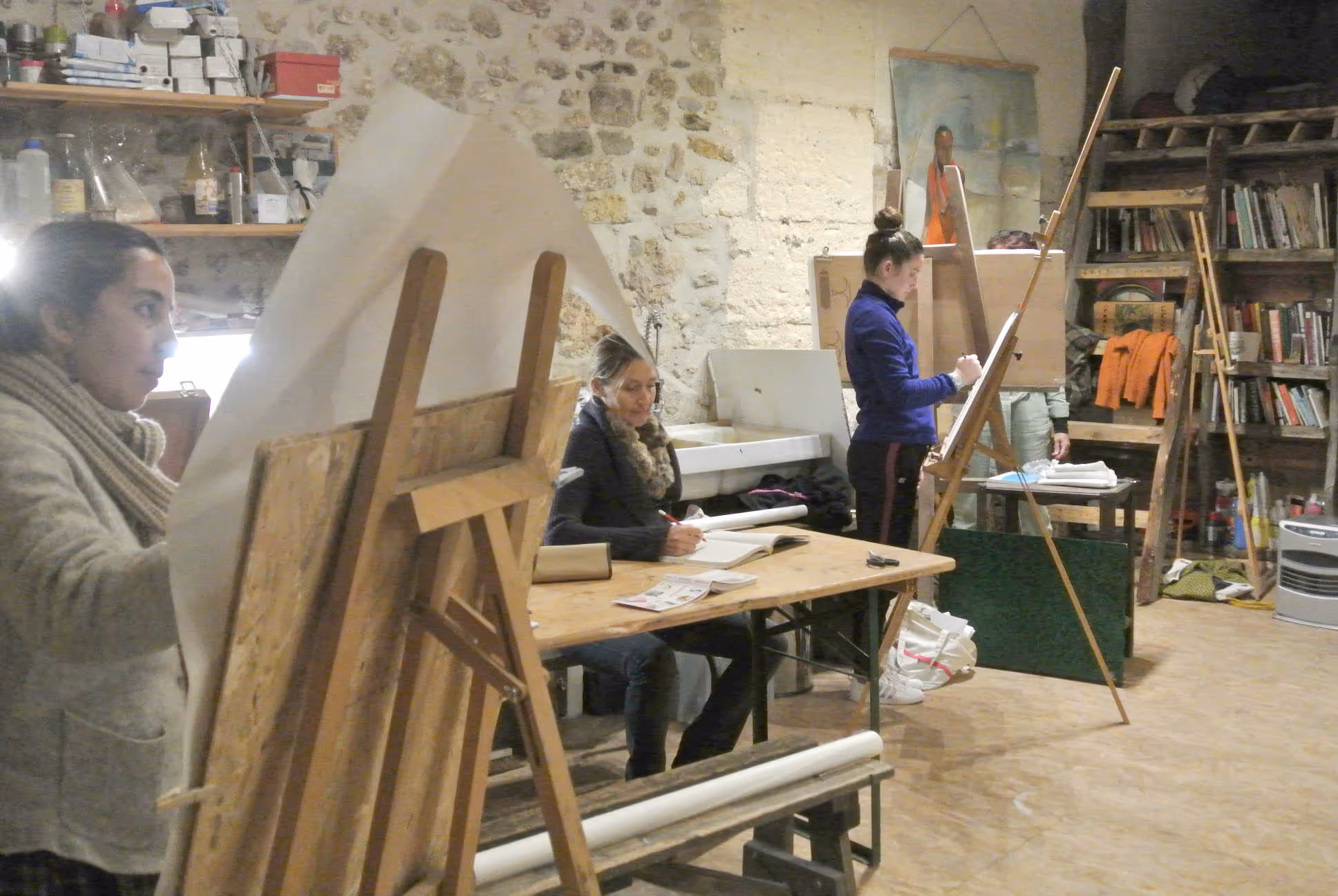 Two women sitting at a table with a laptop and a painting easel.
