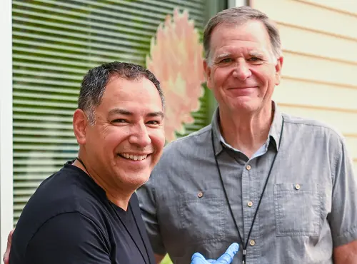 Two smiling middle-aged men standing together outdoors near green blinds