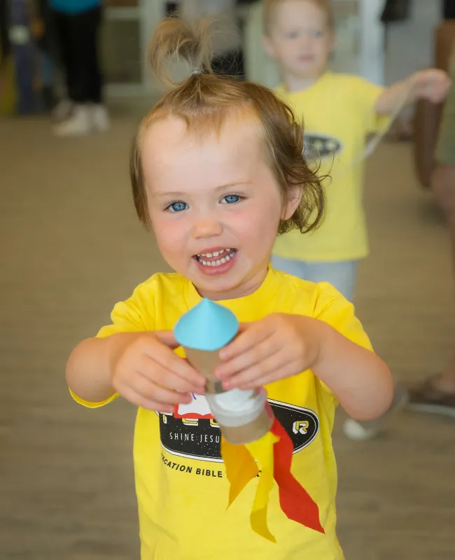 Smiling toddler in yellow shirt holding blue craft item at children's event