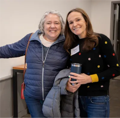 Two women smiling together at an event, wearing warm jackets
