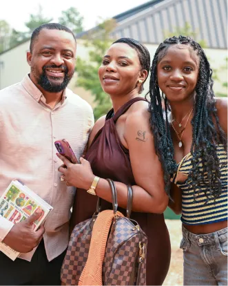 Three people smiling together outdoors, holding phone and book