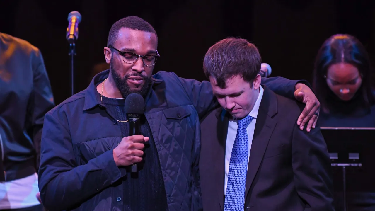 Two men on stage, one speaking into microphone with arm around another