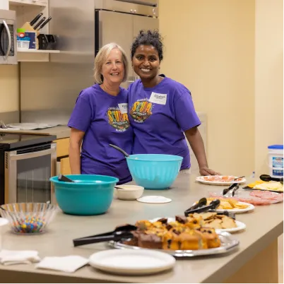 Two volunteers in purple shirts prepare food in a community kitchen