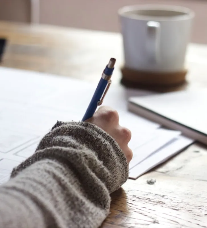 Hand in cozy sweater writing with blue pen near coffee mug