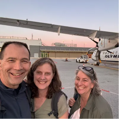 Three smiling friends at airport tarmac with airplane and vehicle in background