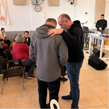 Two men hugging in a community meeting room with people seated nearby