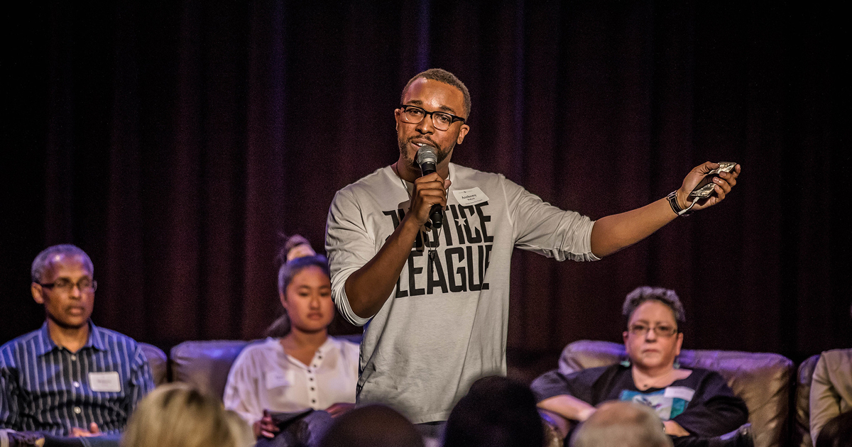 A speaker holds a microphone while addressing an audience at a community event, with people seated on stage behind him.