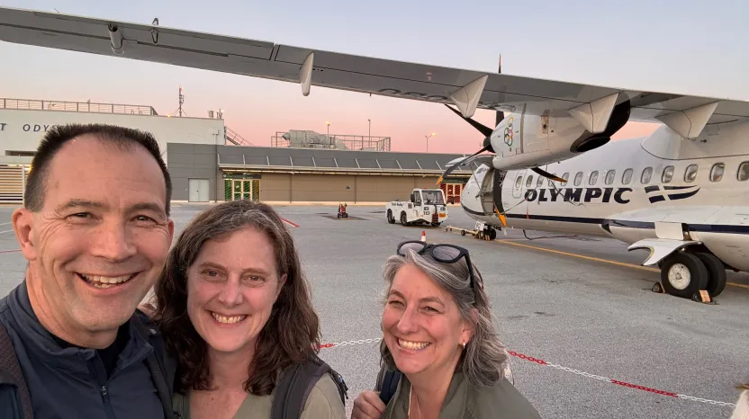 Three smiling travelers at airport with Olympic airplane in background