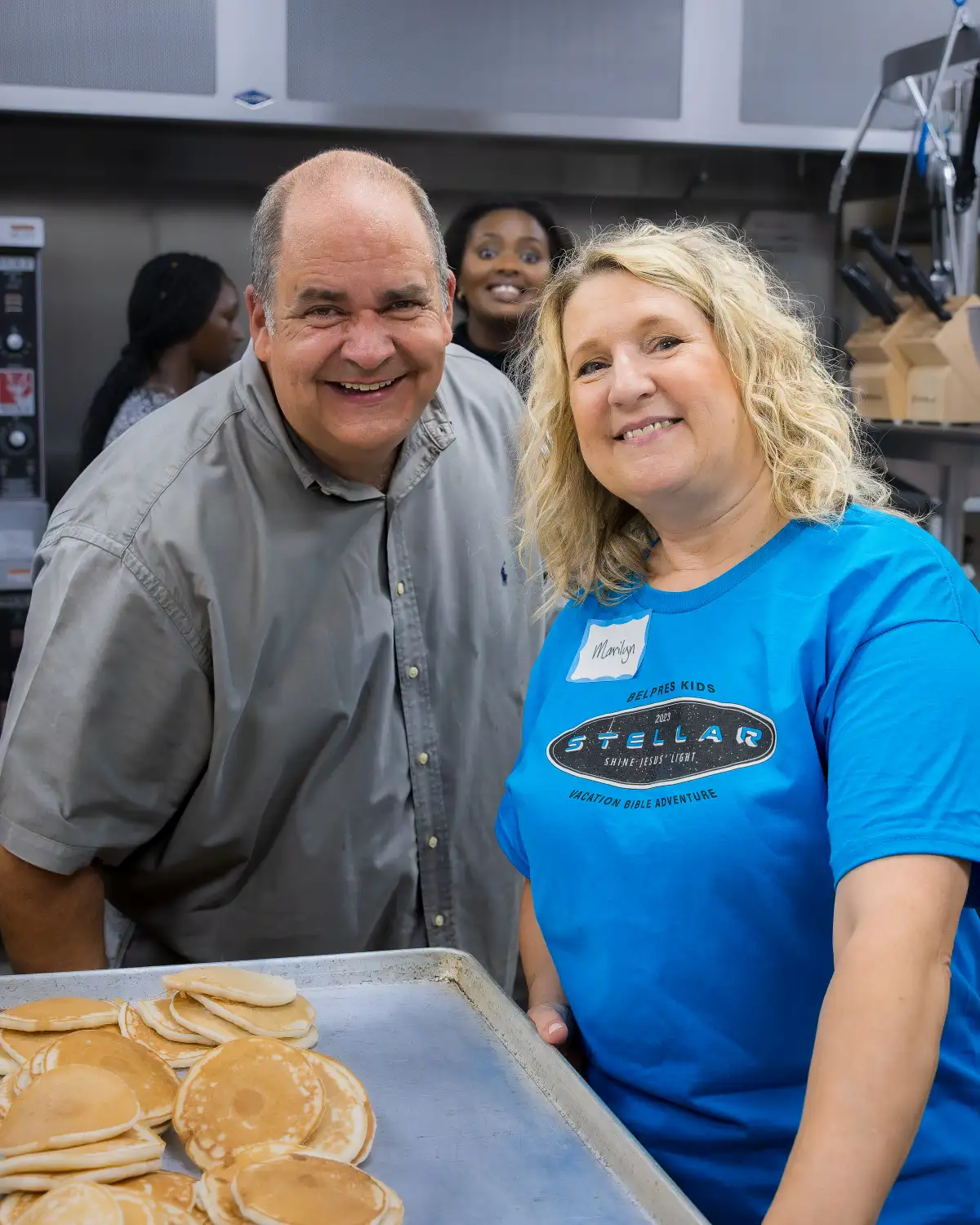 Two smiling volunteers serving pancakes in a kitchen during a community event, with others in the background.
