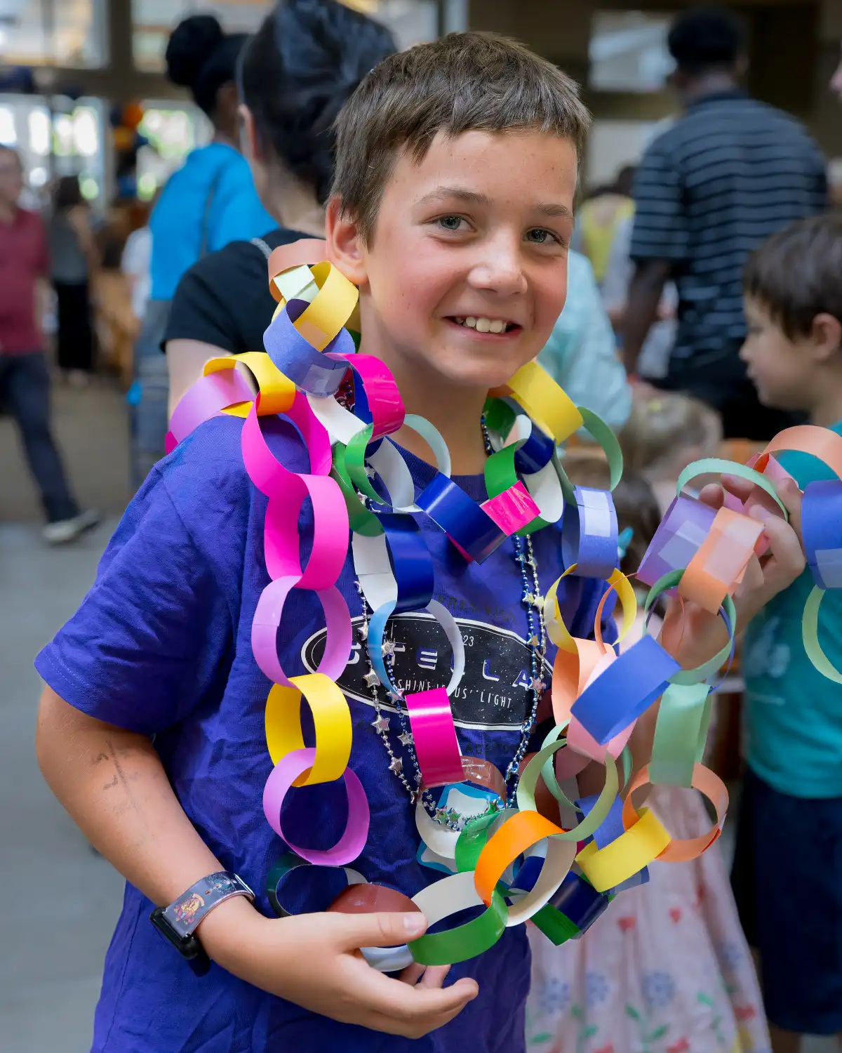Smiling boy wearing a purple shirt and colorful paper chain necklaces during a kids’ craft activity at a community event.