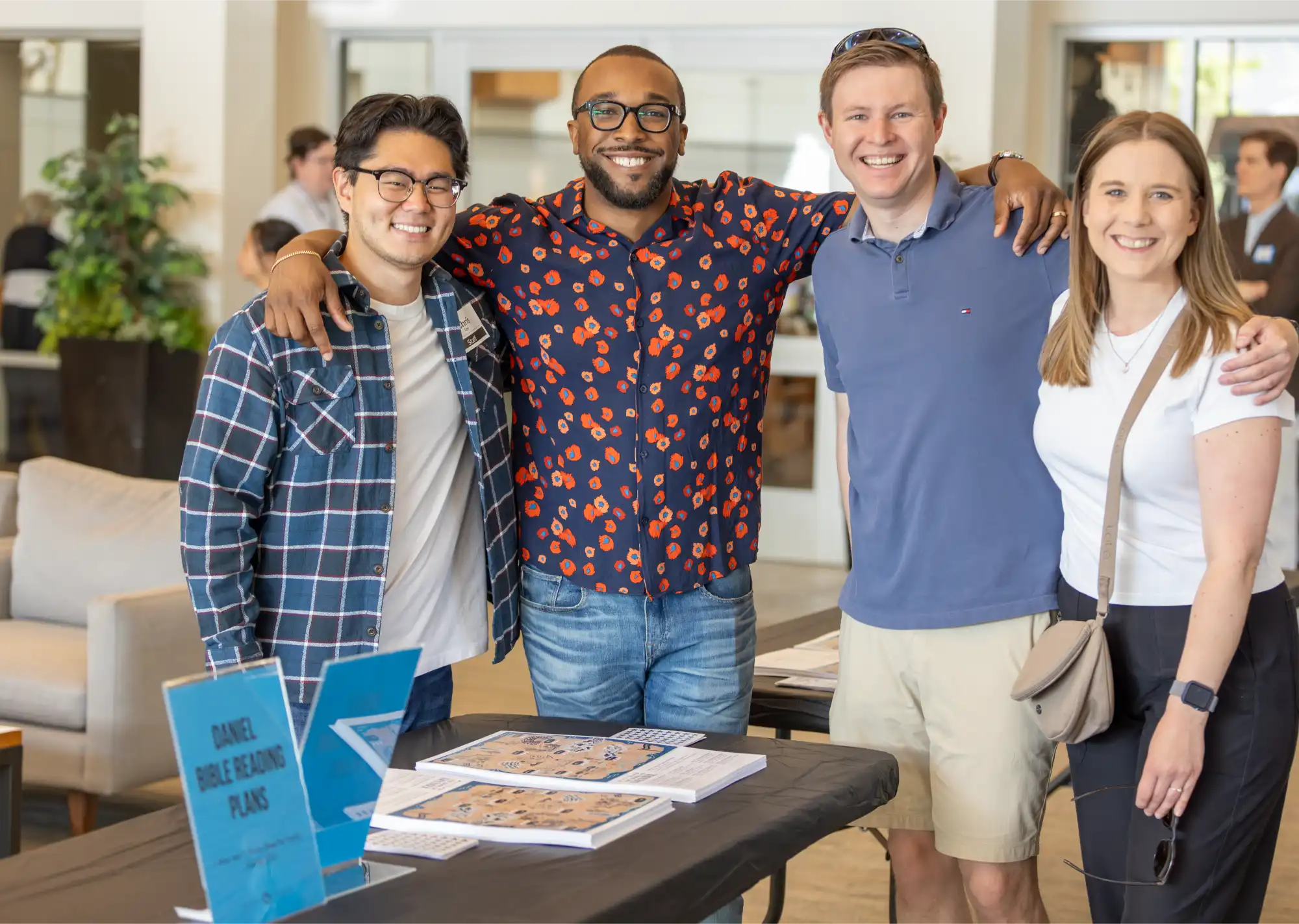 Group of four friends smiling together at a church event table with Bible reading plans displayed in front of them.