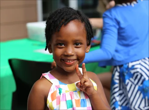 Smiling young girl in a colorful dress holding up a peace sign while standing outdoors at a community event.