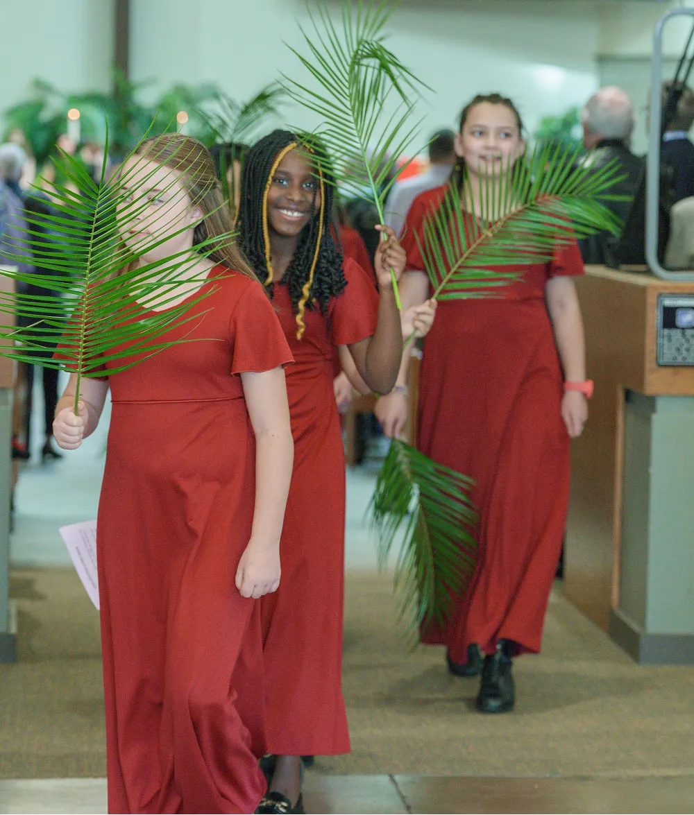 Three girls in red dresses holding palm branches, walking indoors with people in the background.