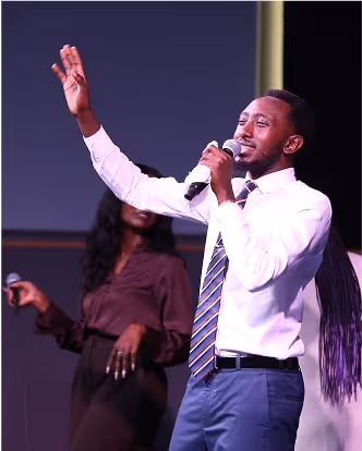 Man in white shirt and tie singing with a microphone and raising his hand on stage.