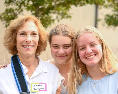 Three smiling females standing closely together outdoors with trees in the background.